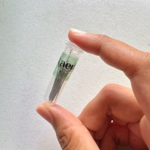A clear tube bottle FILLED WITH NEEDLES with a green label, being held by a hand with a visible fingernail.